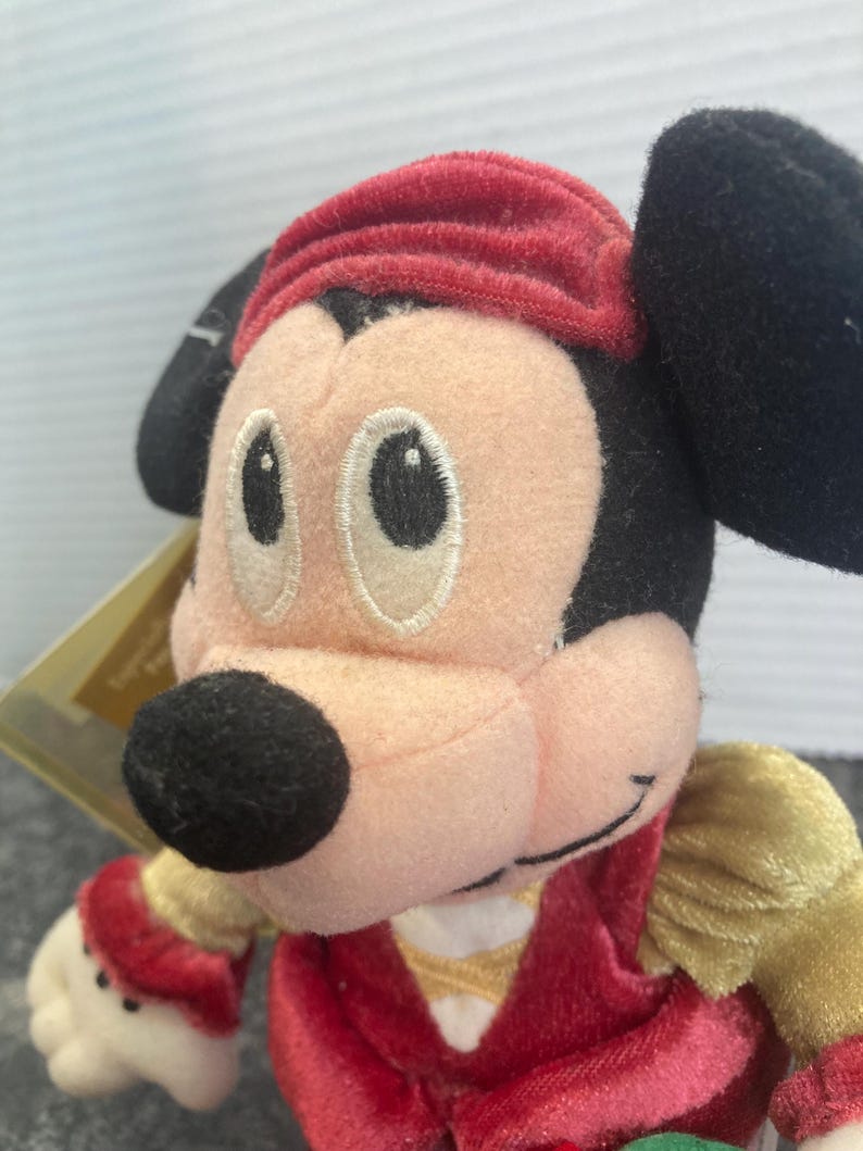May include: A plush Mickey Mouse character toy featuring a peach-colored face, large black ears, and a red velvet-textured hat. The character wears a red velvet vest over gold sleeves, with embroidered eyes and a black nose, capturing a classic look.