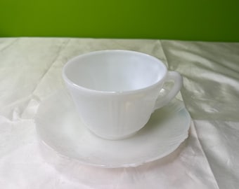 Può includere: Un set di tazza e piattino bianchi. La tazza ha un manico e poggia su un piattino abbinato. Il set è realizzato in materiale bianco lucido e opaco. Lo sfondo è di colore verde.