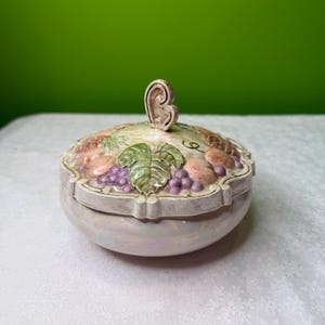 May include: A ceramic trinket box with a lid featuring a relief design of fruits and leaves. The lid has a decorative handle and is adorned with grapes, peaches, and a large leaf. The box has a pearlescent finish.