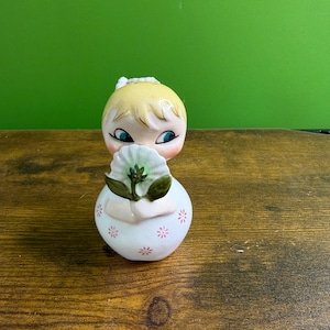 May include: A ceramic figurine of a young girl with blonde hair, blue eyes, and rosy cheeks. She is holding a white flower with green leaves. The figurine is on a wooden surface with a green background.