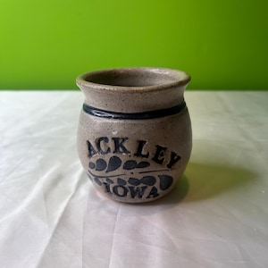 May include: A small, beige ceramic jar with a blue and white design. The jar has the words "Ackley Iowa" written on it.