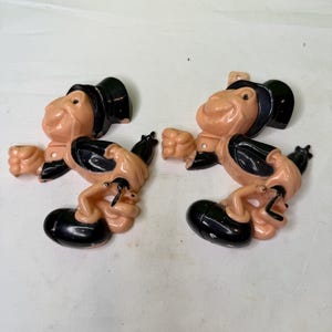 May include: Two vintage, plastic figurines of a cartoon character in a black hat and shoes. The figurines are peach-colored with black accents and appear to be in a dancing pose. The background is a plain white surface.