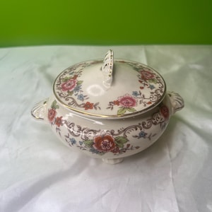May include: Cream-colored ceramic sugar bowl with a lid, adorned with a floral pattern in shades of red, pink, and blue. The bowl has a gold trim and small feet. The lid features a handle for easy lifting.
