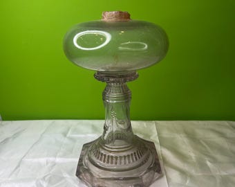 May include: A clear glass oil lamp with a round, wide base and a tall, slender stem. The lamp has a decorative, etched pattern on the stem and base.