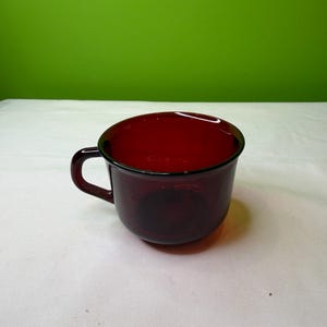 Op de afbeelding: Een donkerrode glazen mok met een afgeronde body en een handvat. De mok is gemaakt van transparant glas. De mok staat op een wit oppervlak tegen een groene achtergrond.