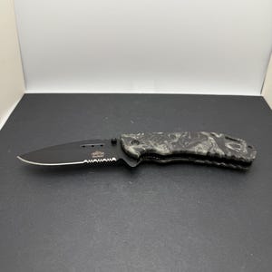 以下が含まれることがあります： 黒い半波刃のブレードとグレーと黒の迷彩柄のハンドルを備えた折りたたみ式ポケットナイフ。ブレードの鋭いエッジの付け根付近には、白とオレンジの文字でMaster USAのロゴが表示されています。