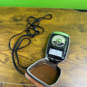 May include: Vintage light meter with a black and green face, housed in a brown leather case with a zipper. A black neck strap is attached. The meter has a dial with numbers and a needle, and a small window with a scale.