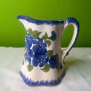 May include: A small white ceramic creamer with a blue and green floral design. The creamer has a handle and a scalloped rim.