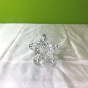 May include: A clear, star-shaped glass candle holder. The candle holder has five points and a small, red accent in the center. The glass is transparent, allowing light to pass through. The background is a white fabric and a green wall.