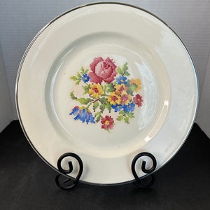 Könnte beinhalten: Cremefarbene Platte mit silbernem Rand, mit einem Kreuzstich-Blumenmuster in Rosa, Gelb, Blau und Grün. Die Platte wird auf einem schwarzen Metallständer präsentiert, was einen Vintage-Look erzeugt.
