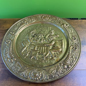 May include: A round, antique brass plate with a detailed relief scene of people gathered around a table. The plate has an ornate, floral border. The plate is a warm, golden color.