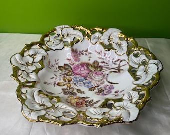 Puede incluir: Un cuenco decorativo de porcelana blanca con un diseño floral. El cuenco tiene un borde festoneado con ribete dorado y diseños de hojas caladas. El interior presenta un ramo de flores rosas, moradas y azules.