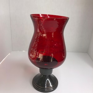 Könnte beinhalten: Ein roter Glas-Kerzenhalter mit einem schwarzen Metallfuß. Der Halter hat einen breiten, runden Fuß und einen hohen, schlanken Körper.