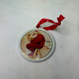 Puede incluir: Adorno navideño redondo con una ilustración de Papá Noel con traje rojo, sentado en un taburete. El adorno tiene un borde dorado y una cinta roja para colgar. La ilustración está firmada "Norman Rockwell".