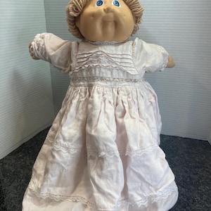 Könnte beinhalten: Eine Puppe mit einem runden, hellbraunen Gesicht, blauen Augen und hellbraunem Wollhaar. Die Puppe trägt ein langes, blassrosa Kleid mit einem hohen Ausschnitt, zarter weißer Spitzenborte, Biesen am Oberteil und gestuften Rüschen.