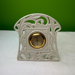May include: Cream-colored ceramic mantel clock with a gold-tone clock face. The clock features an ornate, openwork design with floral and scroll motifs. The clock face has Roman numerals.