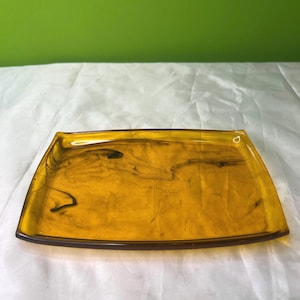 Puede incluir: Una bandeja rectangular de color ámbar con esquinas redondeadas. La bandeja tiene un borde marrón oscuro y un patrón veteado dentro del ámbar. La bandeja está hecha de un material translúcido.