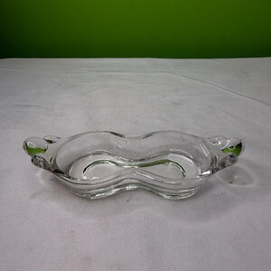 May include: A clear glass serving dish with a unique, wavy design. The dish has two distinct sections, each with a curved shape, and small, rounded handles on either end. The glass is transparent, allowing a view of the background.