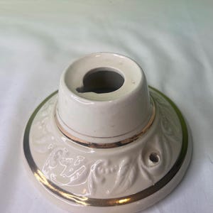May include: White ceramic ceiling fixture with a gold trim and a floral design. The fixture has a hole in the center for a light bulb.