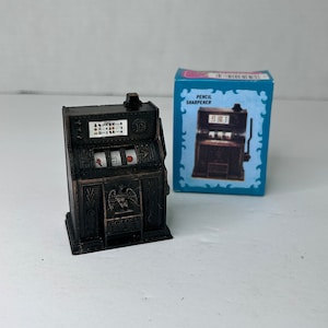 May include: A vintage-style metal pencil sharpener shaped like a slot machine. The sharpener is black with gold accents and features a golden eagle emblem with the words "Golden Eagle" below it. The sharpener has a red, white, and blue color scheme and says "10 Cents" on the side.