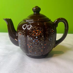 Puede incluir: Una tetera de cerámica marrón con un diseño floral en tonos naranja, azul y dorado. La tetera tiene un cuerpo redondeado y una tapa con un pomo.