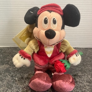 May include: A plush Mickey Mouse toy wearing a red velvet cap, a red vest over gold sleeves, and dark red pants. The character has black ears, a tan face, and white gloves, and is holding a small red fabric rose with green leaves in one hand.