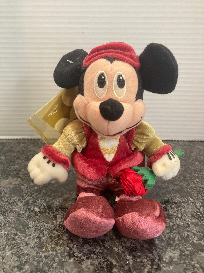May include: A plush Mickey Mouse toy wearing a red velvet cap, a red vest over gold sleeves, and dark red pants. The character has black ears, a tan face, and white gloves, and is holding a small red fabric rose with green leaves in one hand.