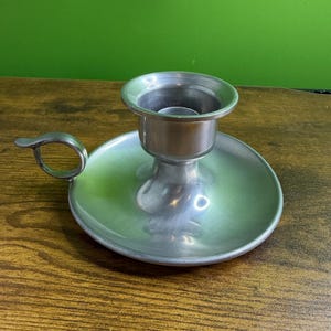 May include: A silver-colored candlestick holder with a circular base and a handle. The candle holder has a cup-shaped top to hold a candle. The handle is a small loop. The background is a green wall and a wooden table.
