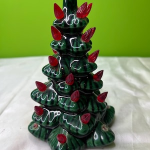 May include: A ceramic Christmas tree with a glossy green finish and red light bulbs. The tree has a classic design with multiple tiers and a pointed top. The lights are evenly spaced throughout the tree, creating a festive look.