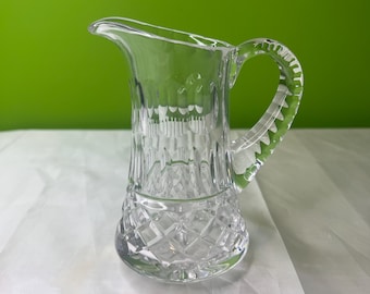 May include: A clear glass creamer with a diamond-cut base and a ribbed body. The handle is also ribbed. The creamer has a spout for easy pouring. The background is a solid green.
