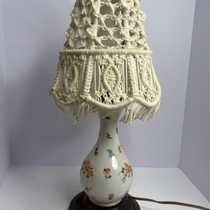 Könnte beinhalten: Vintage Tischlampe mit cremefarbenem Makramee-Lampenschirm und floral gemusterter Keramikbasis. Der weiße Sockel ist mit bunten Blumen und Vögeln verziert. Die Lampe steht auf einem dunkelbraunen Holzsockel.