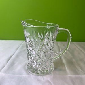 May include: A clear glass pitcher with a diamond pattern. The pitcher has a handle and a wide mouth.