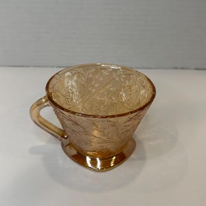 Może przedstawiać: Stara, brzoskwiniowa szklanka z kwiatowym wzorem. Szklanka ma uchwyt i sercowaty spód.