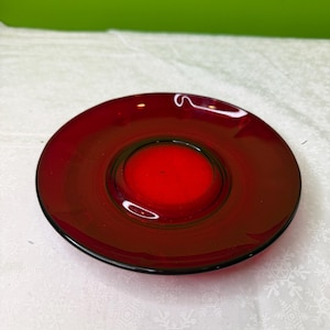 May include: A round, red glass candle holder with a central well. The candle holder has a glossy finish and a dark rim. The background is a white surface with a snowflake pattern.