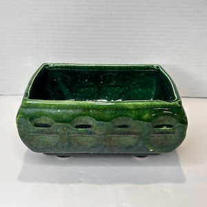 May include: A square green ceramic planter with a textured design and four small feet. The planter is made of glazed pottery and has a dark green colour.