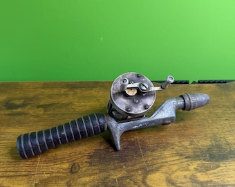 Puede incluir: Carrete de pesca vintage con un mango negro acanalado y un cuerpo de metal plateado. El carrete tiene una manivela y una guía de línea. El carrete está diseñado para ser fijado a una caña de pescar. Fondo de madera y pared verde.