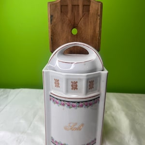 May include: A white porcelain salt cellar with a lid and handle, featuring a floral design and the word "Salt" in gold script. It is mounted on a wooden backing with decorative carvings. The salt cellar is a vintage kitchen item.
