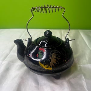 May include: Black ceramic teapot with a rooster design in red, yellow, and gold. The teapot has a metal handle with a spring design and a small lid. The teapot sits on three small feet.