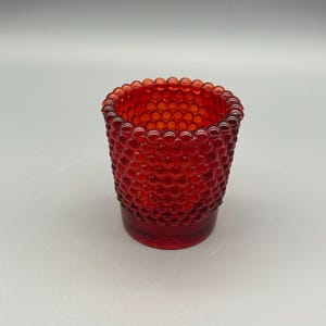 May include: A small, red glass votive candle holder. The exterior features a textured, bubble-like pattern. The rim is rounded, and the base is a solid, smooth red. The candle holder is designed to hold a small candle.