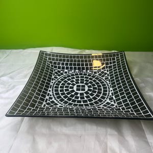 May include: Square black and white decorative plate with a mosaic tile design. The plate features a central circular pattern surrounded by a grid of square tiles. The plate is on a white surface with a green background.