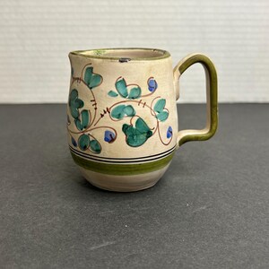May include: A small, white ceramic pitcher with a green and blue floral design. The pitcher has a handle and a rounded body.