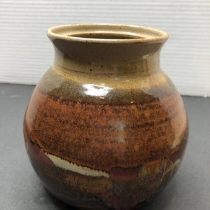 May include: A round, brown ceramic vase with a wide mouth and a mottled glaze. The glaze features shades of brown, orange, and white.