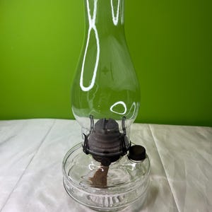 Puede incluir: Una l&aacute;mpara de aceite de vidrio transparente con una base cil&iacute;ndrica y una chimenea alta y curvada. La l&aacute;mpara presenta un conjunto de quemador de metal oscuro y una mecha. La l&aacute;mpara est&aacute; dise&ntilde;ada para contener aceite y proporcionar luz.