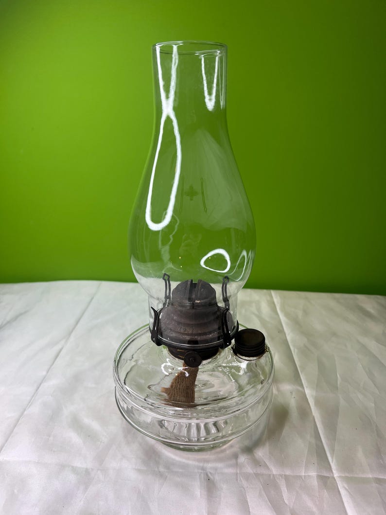 Puede incluir: Una l&aacute;mpara de aceite de vidrio transparente con una base cil&iacute;ndrica y una chimenea alta y curvada. La l&aacute;mpara presenta un conjunto de quemador de metal oscuro y una mecha. La l&aacute;mpara est&aacute; dise&ntilde;ada para contener aceite y proporcionar luz.