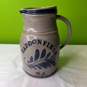 Puede incluir: Una jarra de gres gris con un esmalte azul y un diseño azul de una hoja y las palabras "Haddonfield, N.J."