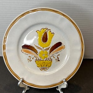Puede incluir: Un plato de cerámica blanco con un borde marrón y un diseño floral en tonos amarillos, marrones y naranjas. El diseño presenta un tulipán, una flor más pequeña y una maceta marrón con un borde amarillo.