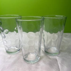 Op de afbeelding: Drie heldere drinkglazen met een getextureerd, gestippeld patroon. De glazen zijn gemaakt van transparant glas en zijn leeg. De achtergrond is effen groen en de glazen staan op een wit oppervlak.