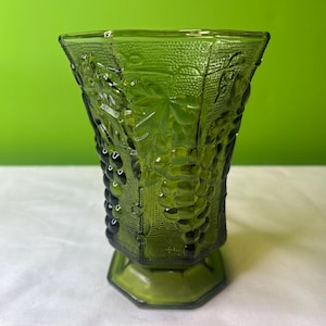 May include: A green glass vase with a grapevine design. The vase has a textured surface and a wide base.