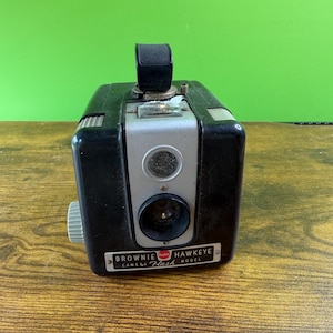 May include: A vintage black Kodak Brownie Hawkeye Flash camera. The camera has a silver face with a lens and flash. The camera has a black handle on top and a white dial on the side. The camera is sitting on a wooden surface.