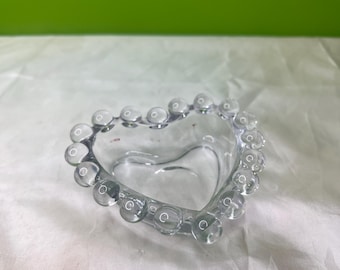 Peut inclure: Un plat en forme de cœur en verre transparent avec un bord perlé. Le plat est transparent, laissant passer la lumière, et repose sur une surface blanche avec un fond vert.